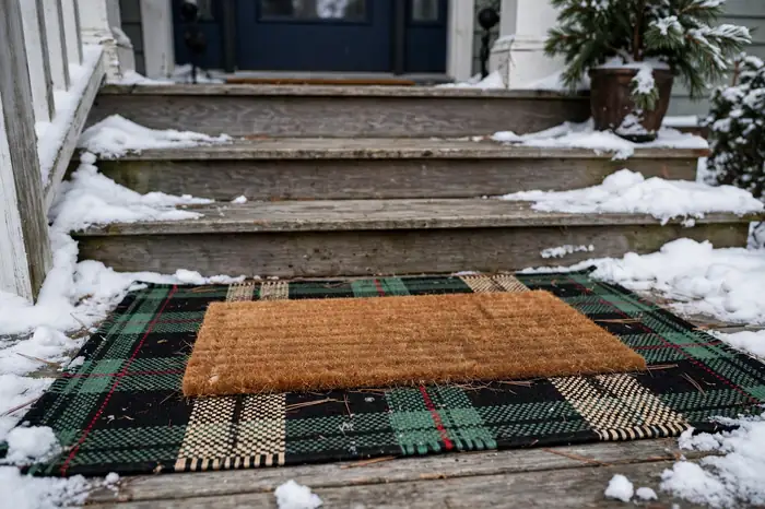 Photorealistic photograph of a layered welcome mat over a plaid outdoor rug on a winter front porch